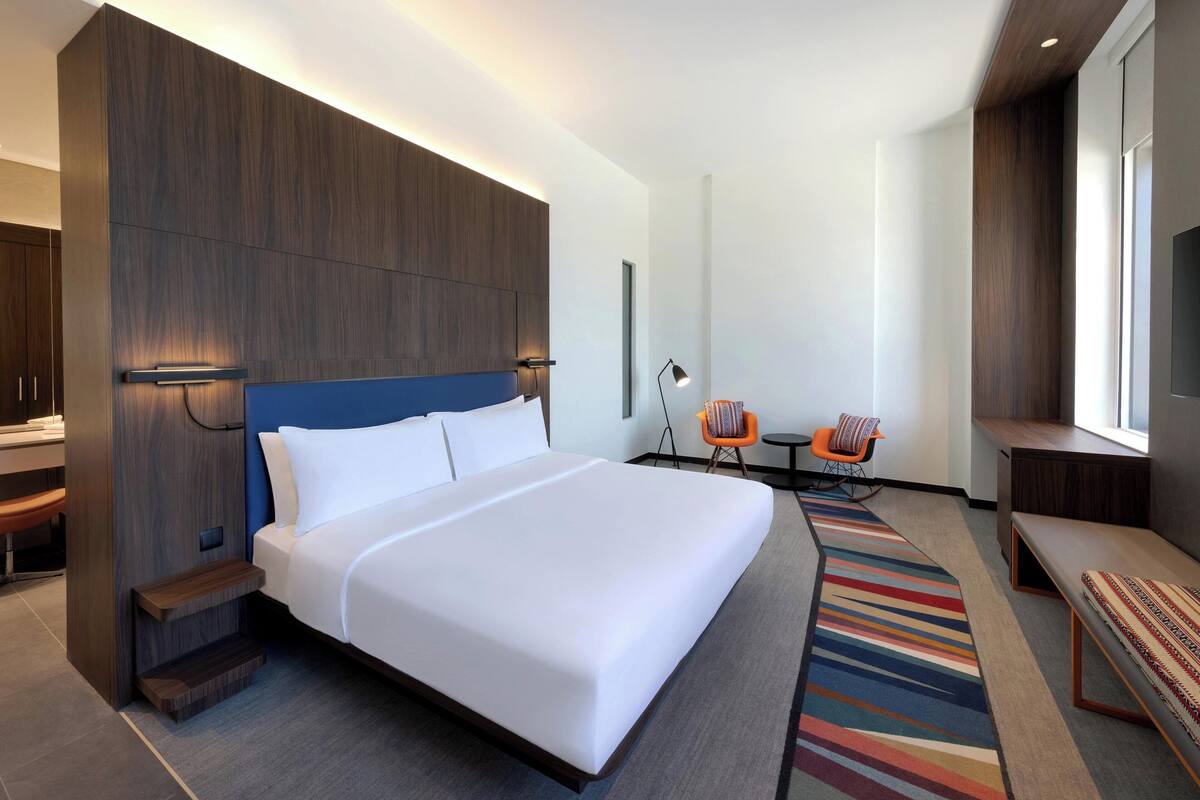 A modern hotel room with a large bed, a desk, a chair, and a bench.