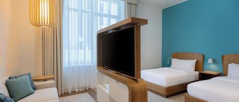 Une chambre d’hôtel avec une télévision posée sur un meuble en bois, deux lits, un canapé et une fenêtre avec des rideaux.