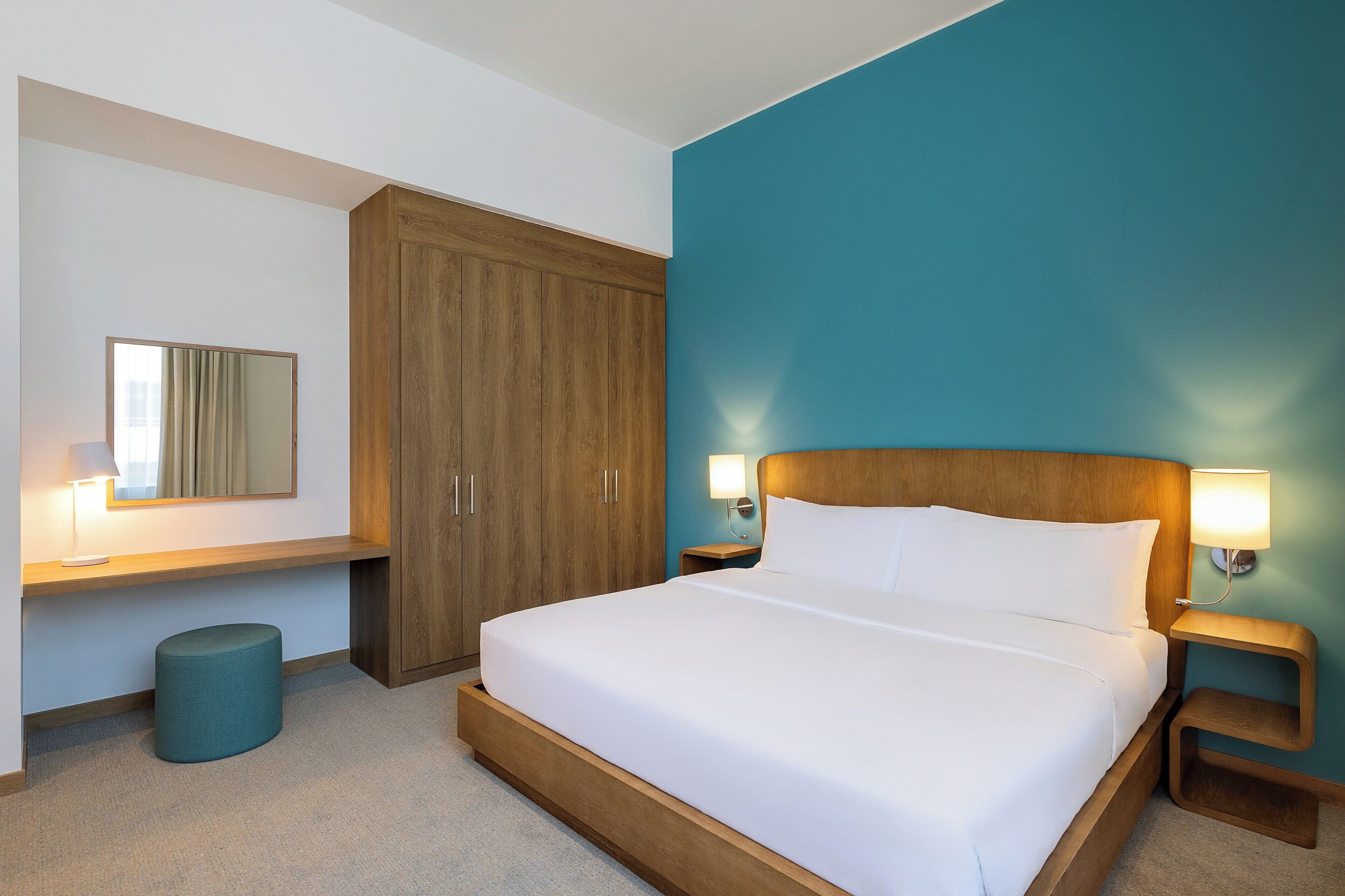 Una habitación de hotel con una cama grande, un armario de madera, dos mesitas de noche con lámparas, un otoman redondo y un espejo en la pared.