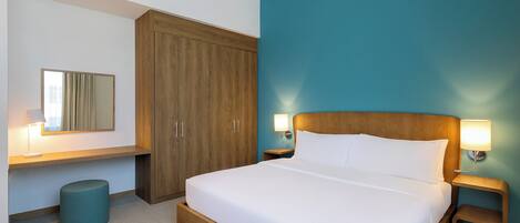 A hotel room with a large bed, wooden wardrobe, two bedside tables with lamps, a round ottoman, and a mirror on the wall.