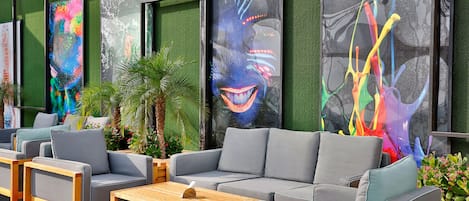 Un espacio exterior moderno con sofás grises y mesas de madera, enmarcado por colorida obra de arte abstracta.