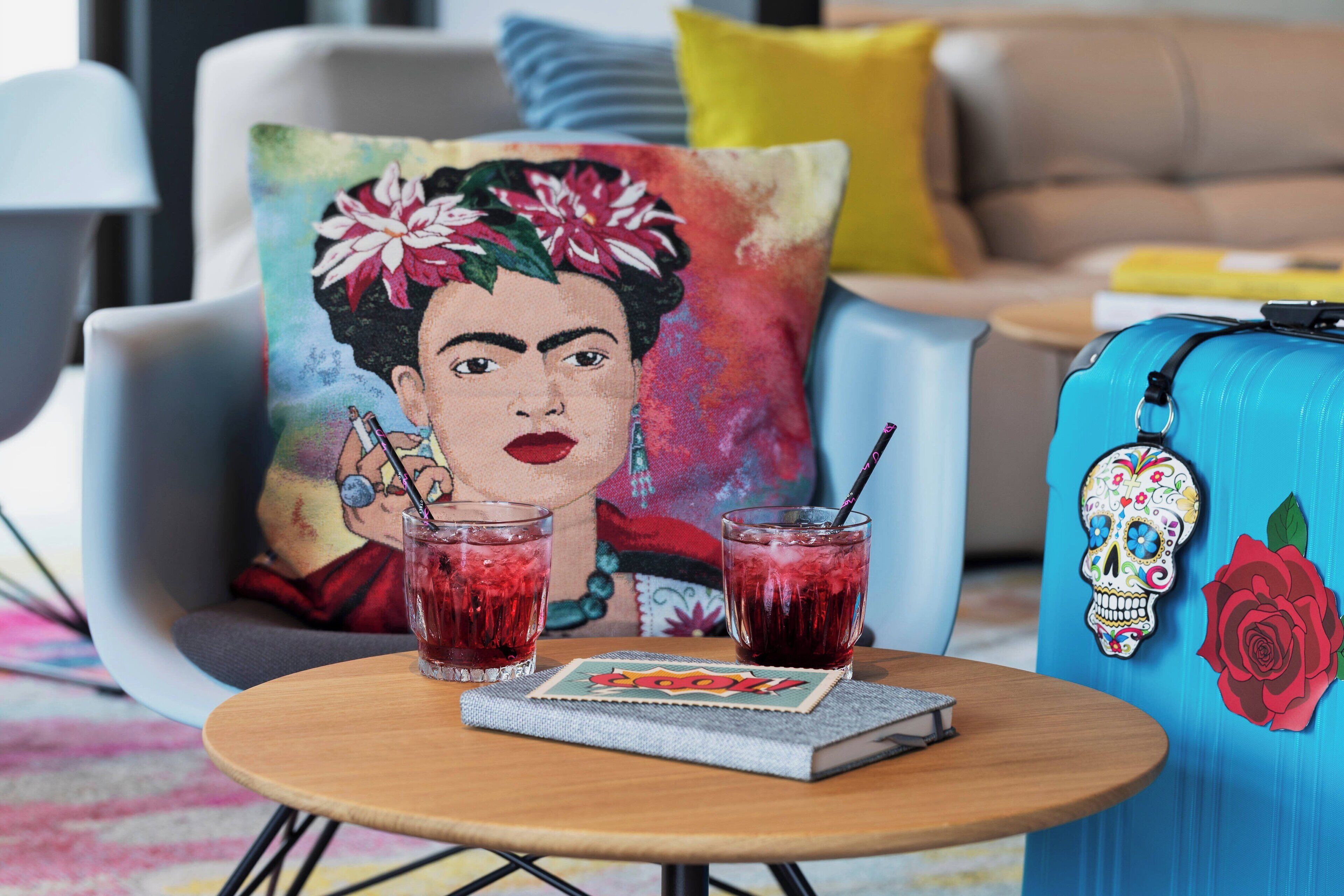 A cozy seating area with a colorful pillow, a blue suitcase, two glasses of red beverage, and a notebook on a small round table.