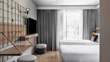 A modern hotel room with a TV mounted on a wall, a wooden floor, two beds, a small round ottoman, and a window with curtains.