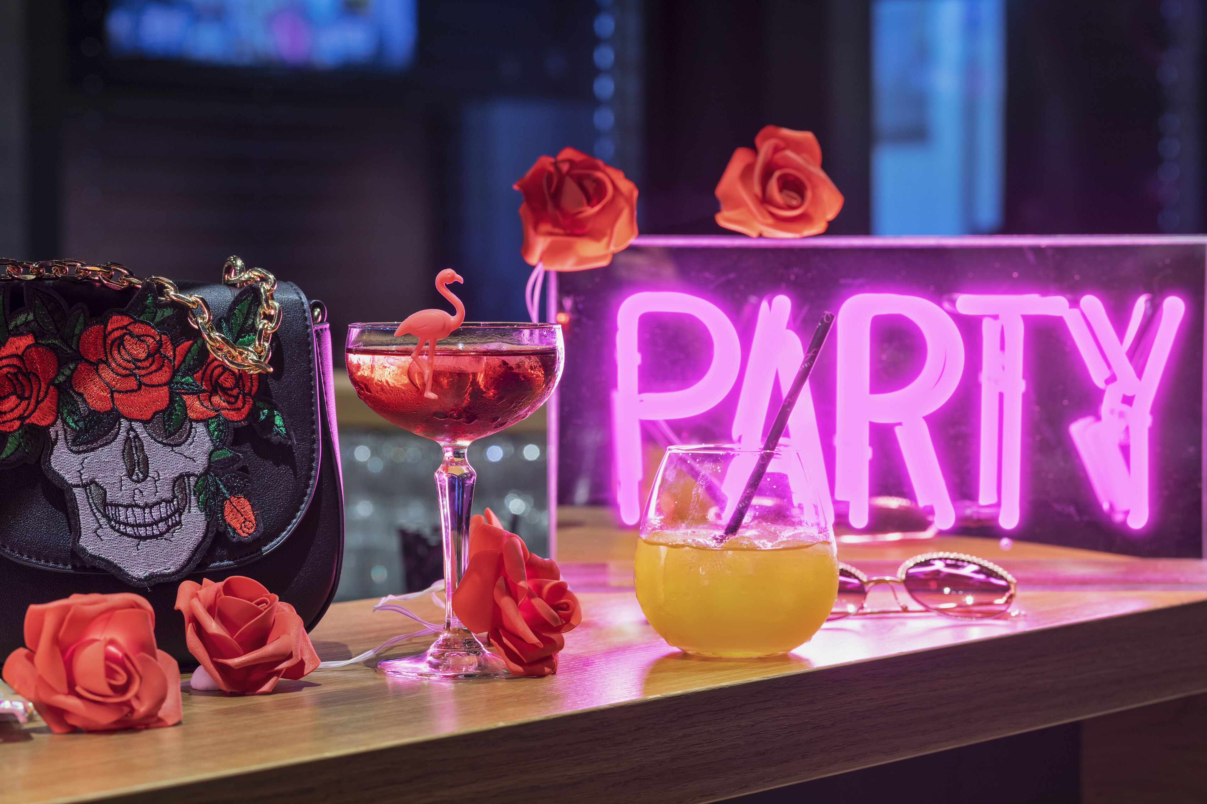 A bar area with a neon sign, a cocktail, a skull-themed bag, and rose decorations.