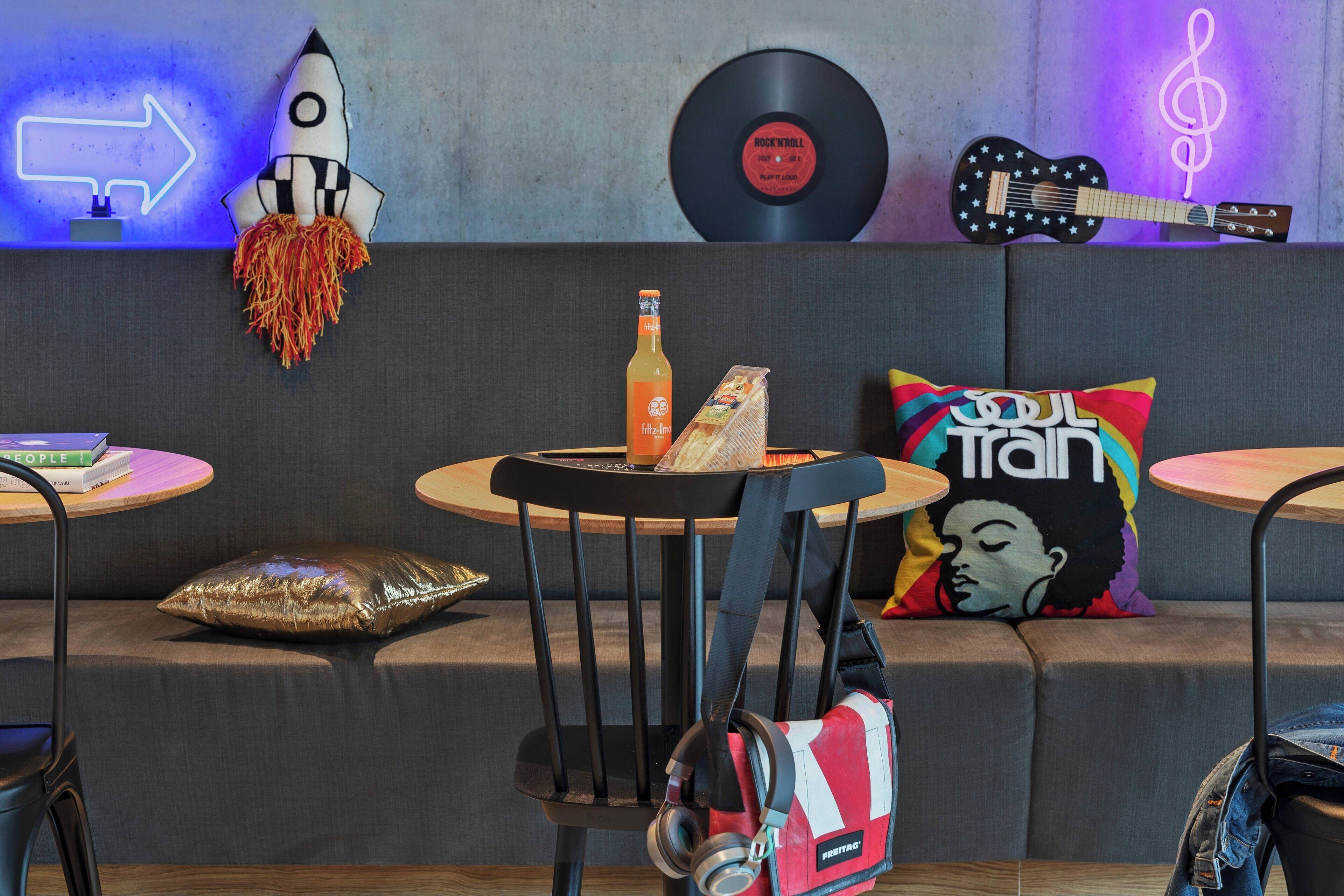 Un área de descanso moderna con un sofá, dos mesas redondas pequeñas, una decoración con forma de cohete, un disco de vinilo, una guitarra y un letrero de neón.
