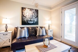 A room with a dark leather sofa, striped pillows, a large tiger painting, two bedside tables with lamps, and a large window with blinds.