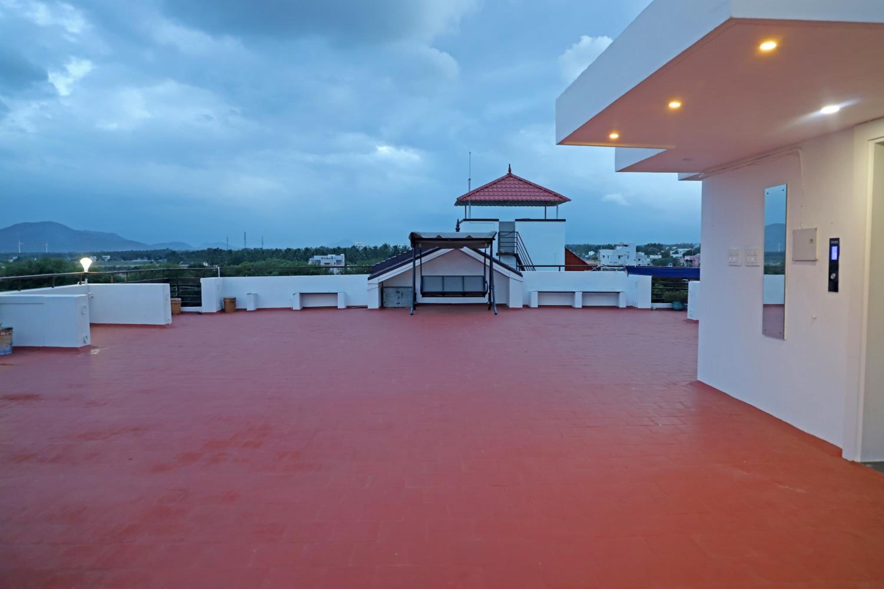 A rooftop terrace with a red floor, a covered seating area, and a view of distant buildings and mountains.