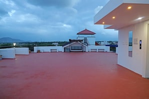 Une terrasse sur le toit avec un sol rouge, un espace salon couvert et une vue sur des bâtiments et des montagnes situés au loin.