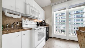 A modern kitchen with white cabinets, a granite countertop, a microwave, stove, and a refrigerator. It features a large window with a view of a multi-story building.