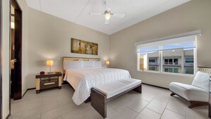 A bedroom with a bed, bedside tables, a chair, a ceiling fan, a window, and a painting on the wall.