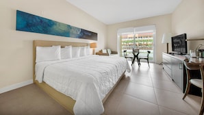 A hotel room with a large bed, a desk with a TV, and a balcony with a view of palm trees.