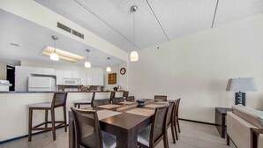 A modern kitchen with white cabinets, a microwave, and a sink. There is a dining area with a large table and chairs, and a lamp on a side table. - Perfect Fully Furnished 3Bedroom Condo Ocean View  (South Padre Island)