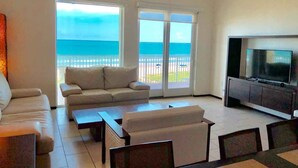 A living room with a sofa, coffee table, TV, and a balcony with a view of the ocean. - Perfect Fully Furnished 3Bedroom Condo Ocean View  (South Padre Island)