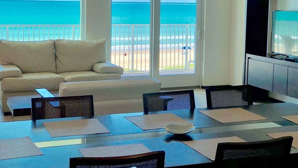 A room with a large window overlooking a beach, a sofa, a dining table with chairs, and a TV. - Perfect Fully Furnished 3Bedroom Condo Ocean View  (South Padre Island)