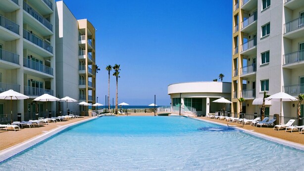 A hotel pool area with sun loungers and umbrellas, surrounded by multi-story buildings with balconies.