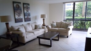 A living room with two sofas, a coffee table, and a lamp. There are paintings on the wall and a window with a view of greenery. - Luxurious Beach Front 1st Floor 2/2 Condo (Fort Pierce)