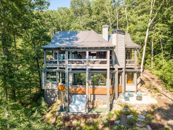 Une maison en bois à deux étages, dotée d’un jacuzzi et d’un espace extérieur aménagé, le tout entouré d’arbres.