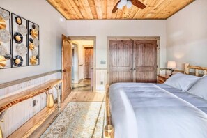 A bedroom with a wooden ceiling, a large bed, a nightstand, a dresser, and a hallway leading to another room.