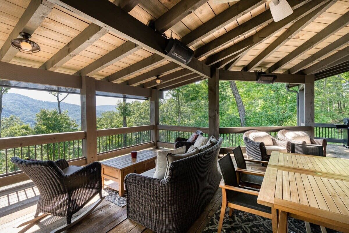 A covered outdoor deck with wicker furniture, wooden tables, and a view of the mountains.