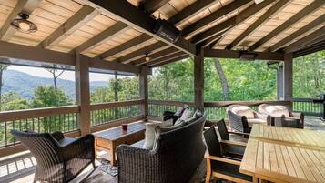 A covered outdoor deck with wicker furniture, wooden tables, and a view of the mountains.