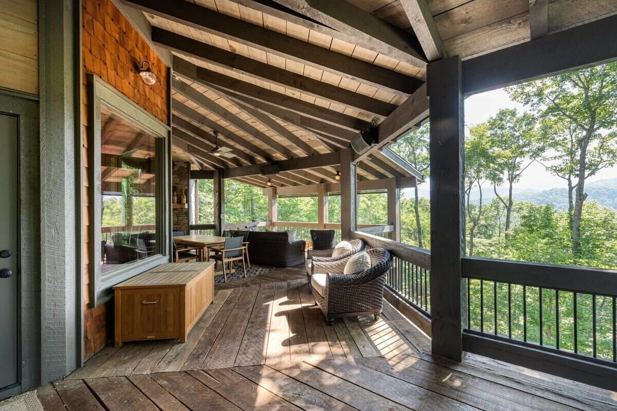 A covered outdoor area with wooden flooring, a seating area with a table and chairs, and a view of the surrounding forest.