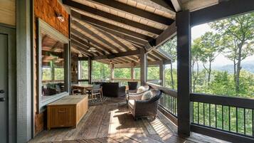 A covered outdoor area with wooden flooring, a seating area with a table and chairs, and a view of the surrounding forest.