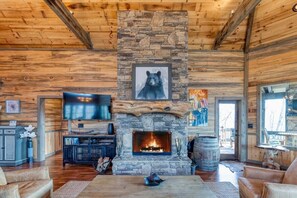 A rustic cabin interior with a stone fireplace, wooden walls, a mounted bear picture, a TV, and a dining area.