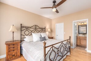 Un dormitorio con una cama de madera, dos mesitas de noche con lámparas, un ventilador de techo y una puerta que da a un baño con espejo y lavamanos.