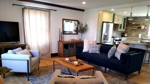 A modern living room with a sofa, coffee table, and TV. It includes a kitchen area with a refrigerator and cabinets.