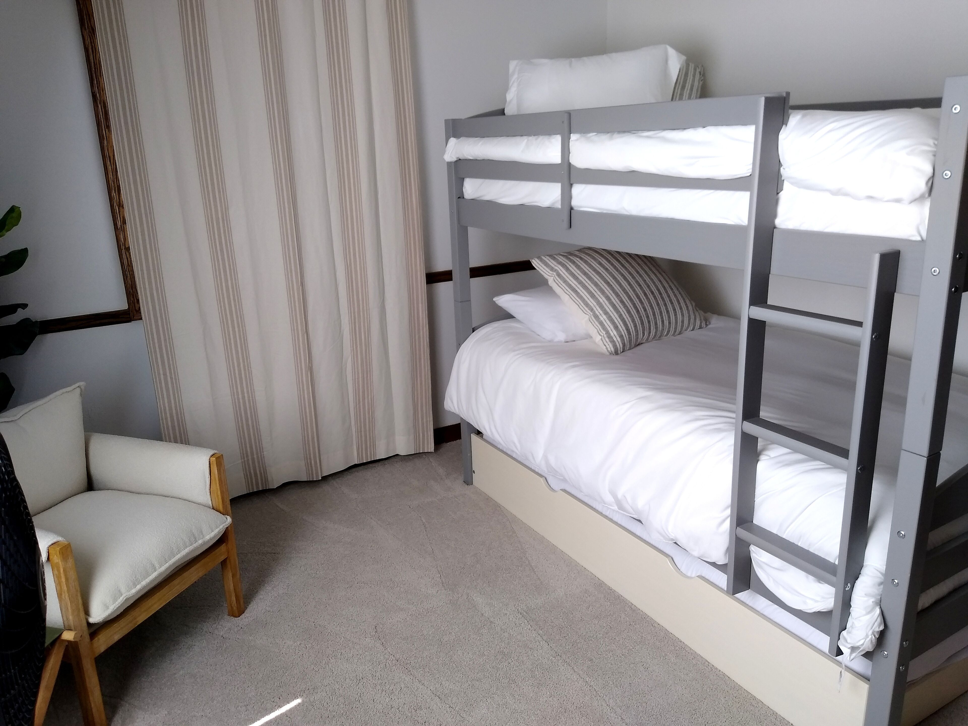 Un lit superposé avec une literie blanche et des oreillers à rayures, une chaise en bois et un rideau à rayures.