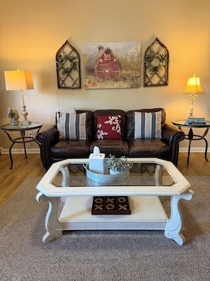 A living room with a brown leather sofa, a white coffee table with a tissue box and decorative items, and two wall-mounted lamps. - Brick Farmhouse Retreat – 7 Minutes to Red Clay State Park (Cleveland)