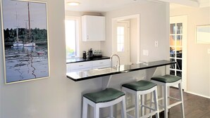 A modern kitchen with a black countertop, white cabinets, and three bar stools with green cushions. - Cape Gem in New Seabury resort & near the beach. (Mashpee)