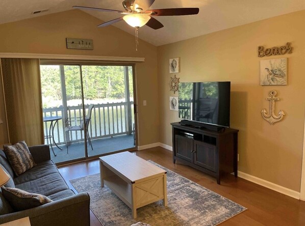 A living room with a sofa, coffee table, TV, and a balcony with outdoor furniture. - Pawleys Paradise (Pawleys Island)