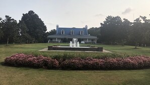 Un grand bâtiment imposant, avec une fontaine en premier plan, entouré de pelouses bien entretenues et d’arbustes en fleurs.