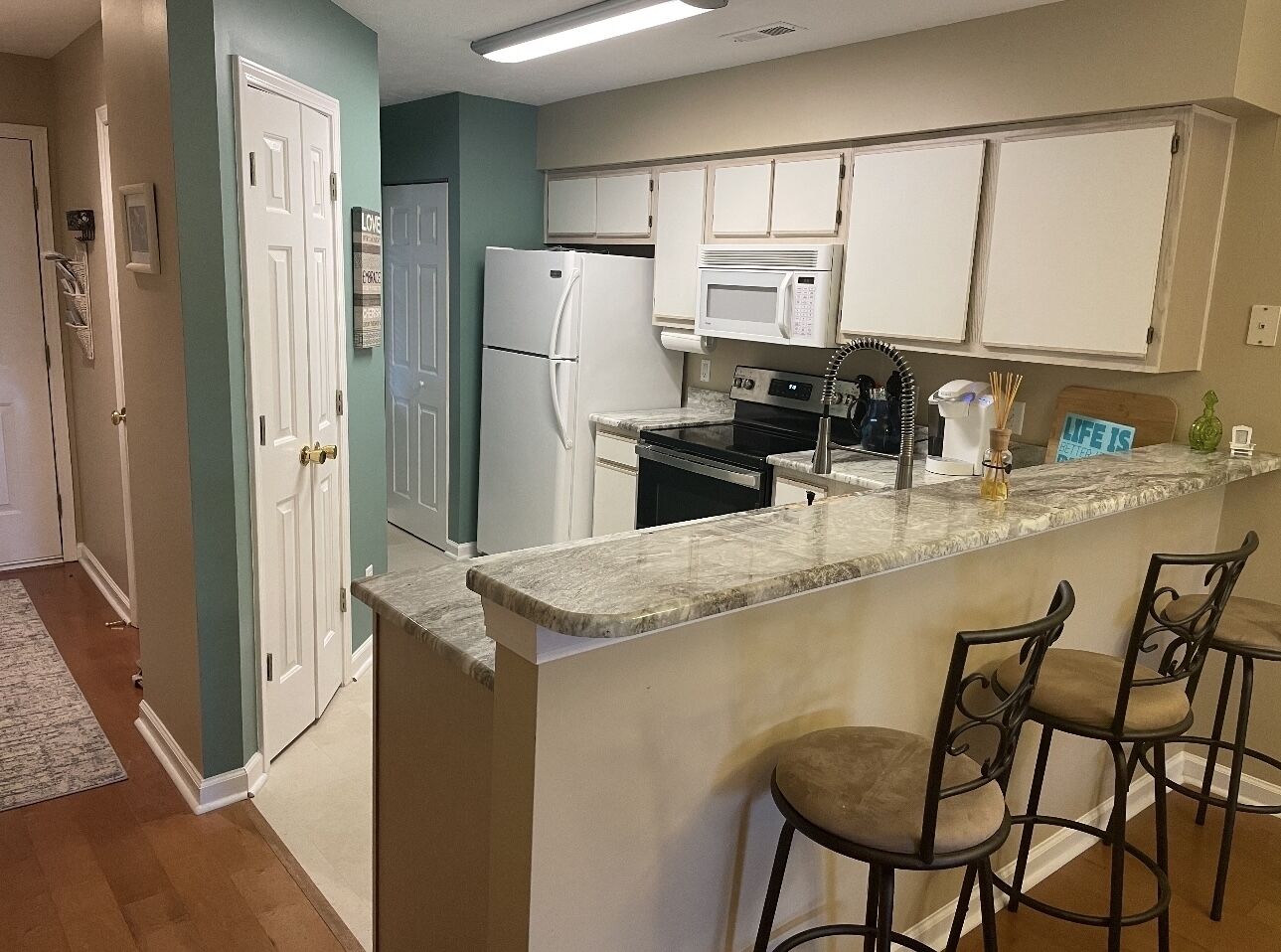 A kitchen with white cabinets, a refrigerator, a microwave, and a stove. There are three bar stools and a small island with a marble countertop.