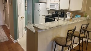 Une cuisine avec des meubles blancs, un réfrigérateur, un four à micro-ondes et une cuisinière. On y trouve trois tabourets de bar et un petit îlot central doté d’un plan de travail en marbre.