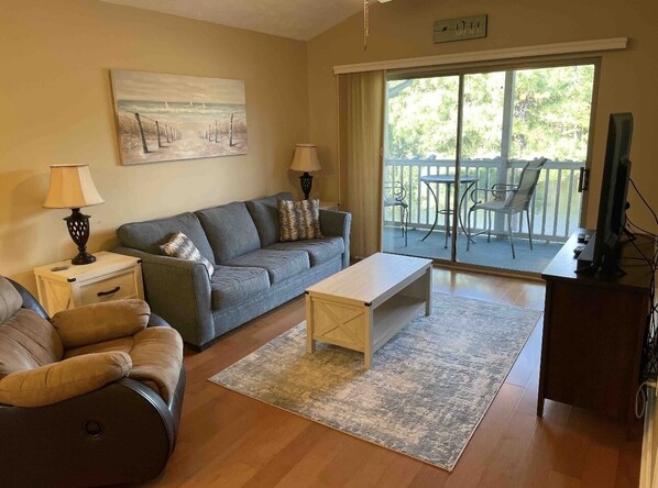 A living room with a sofa, coffee table, and a TV. There is a balcony with chairs and a table.