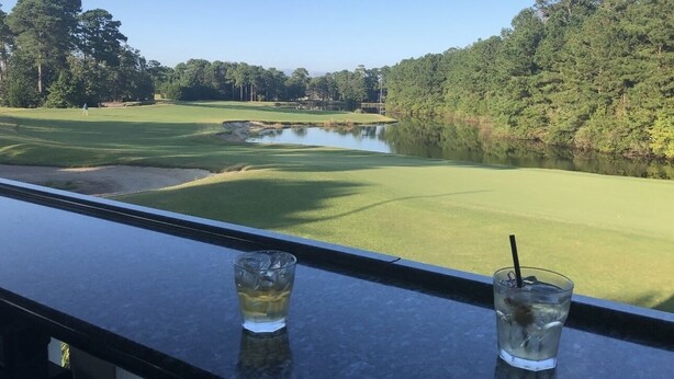 Une vue depuis un balcon donnant sur un terrain de golf avec un étang, et deux verres contenant une boisson posés sur la balustrade.