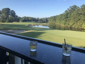 A view from a balcony overlooking a golf course with a pond, with two glasses of a beverage on the railing. - Pawleys Paradise (Pawleys Island)