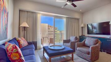 Habitación de hotel con sofá, sillón, mesa de centro, televisor y balcón con vistas a edificios.