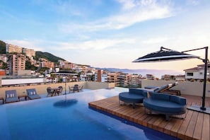 A rooftop infinity pool with lounge chairs and a wooden deck, overlooking a cityscape with buildings and hills. - Top-Floor Penthouse · Walk to Beach · King Bed (Puerto Vallarta)