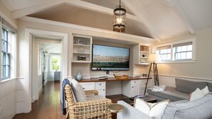 A spacious living area with a large flat-screen TV, a wicker chair, a grey sofa, and a wooden dining table.