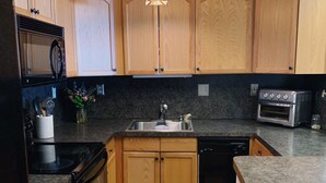 A modern kitchen with wooden cabinets, a black countertop, a built-in oven, and a unique pendant light.
