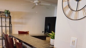 A modern kitchen with a central island, wooden chairs, a ceiling fan, and a decorative clock on the wall.
