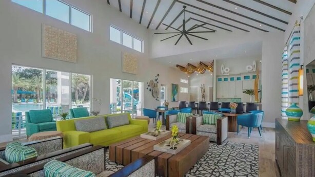 A spacious living area with a bright yellow sofa, a wooden coffee table, and blue chairs. There are large windows providing natural light and a view of the outdoors. - Lux Disney Villa | Pool, Theater/games | Sleeps 22 (Kissimmee)