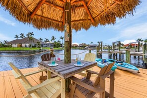 Une terrasse en bois surmontée d’un parasol en chaume, une table avec des chaises, et une vue sur une étendue d’eau où l’on aperçoit des bateaux et des maisons au loin.