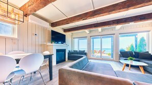A spacious living area with a wooden ceiling, a fireplace, and large windows offering a view of the outdoors.