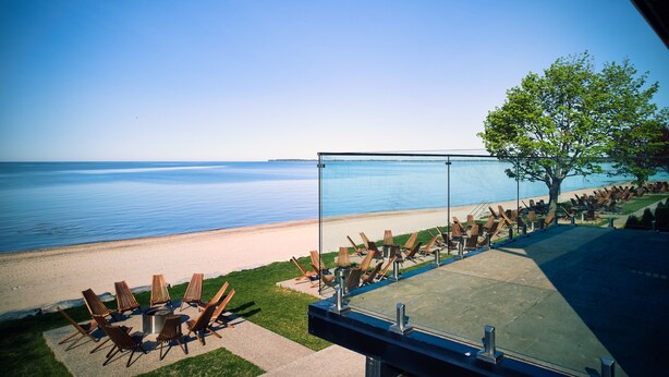 A beachside seating area with lounge chairs and a fire pit, overlooking a calm sea and a clear sky.