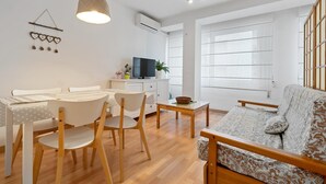 A modern living room with a sofa, dining table, and chairs. There is a television on a cabinet, and a hanging light fixture.