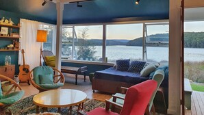 A cozy living room with a large window overlooking a lake, furnished with a sofa, chairs, and a small table. - Reimagined Designer Lakehouse in Prime Location (Burrill Lake)